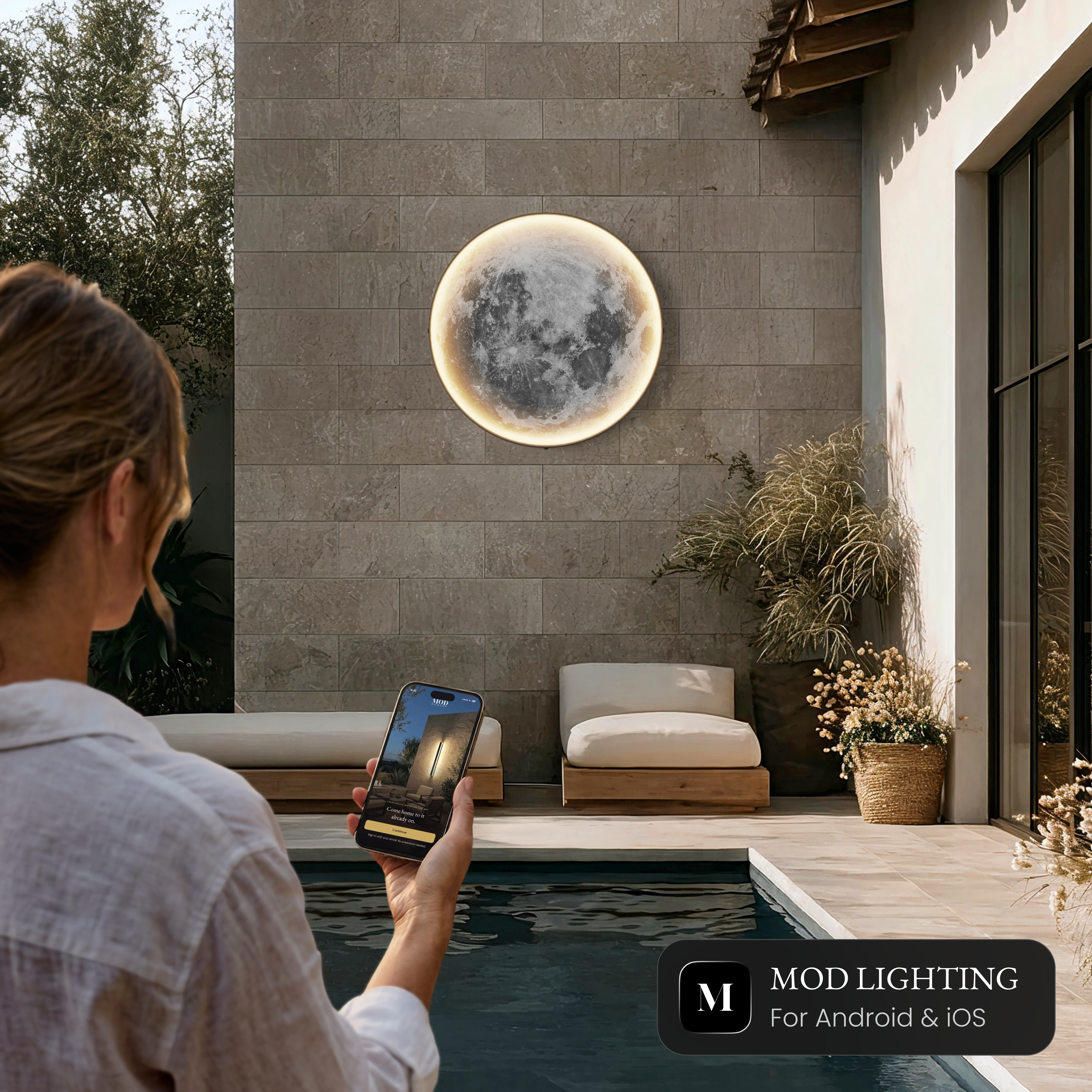 Luna Outdoor Smart moon wall light beside a pool, controlled from the MOD LIGHTING app on a phone