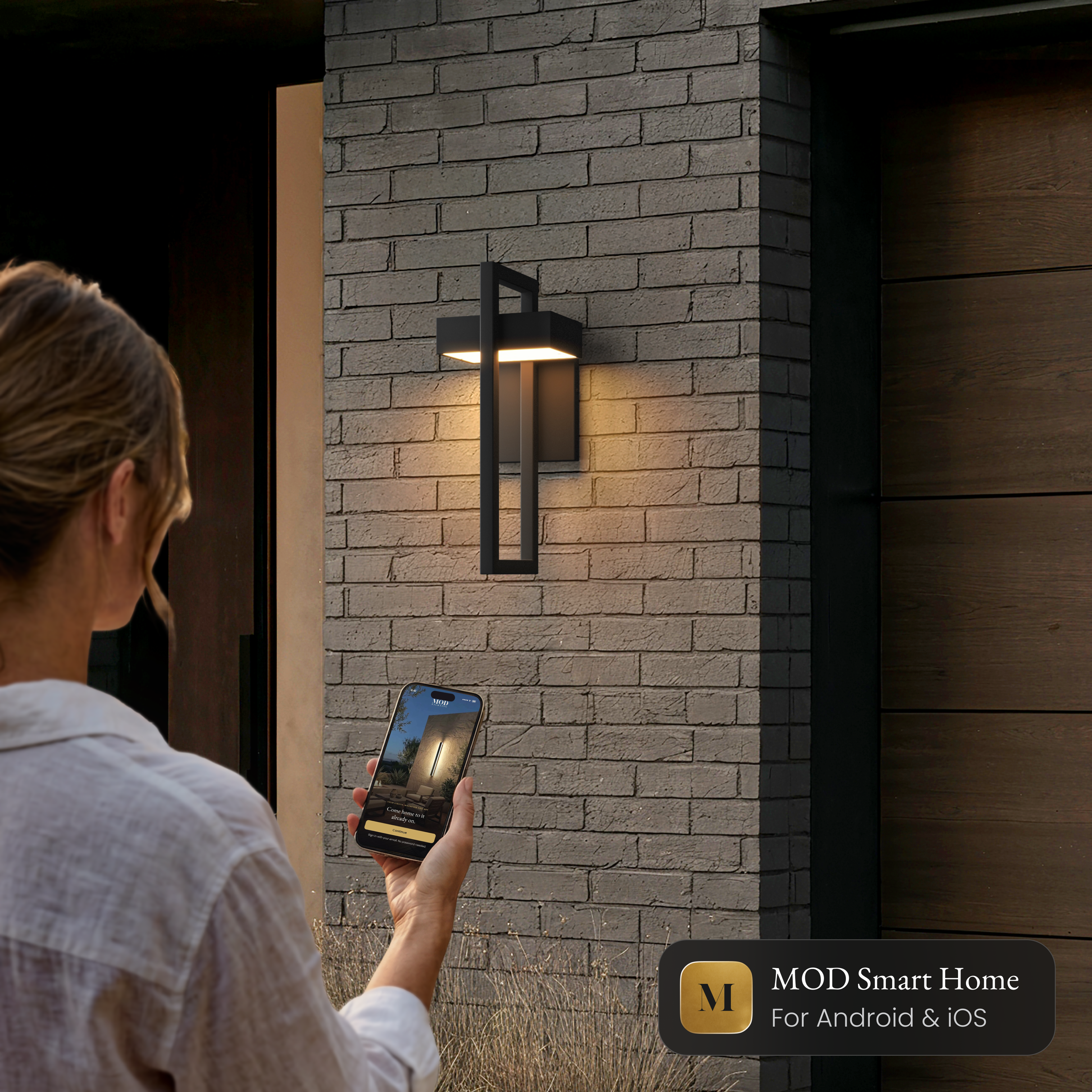 Terri Smart black outdoor sconce on a brick wall, controlled from the MOD Smart Home app on a phone