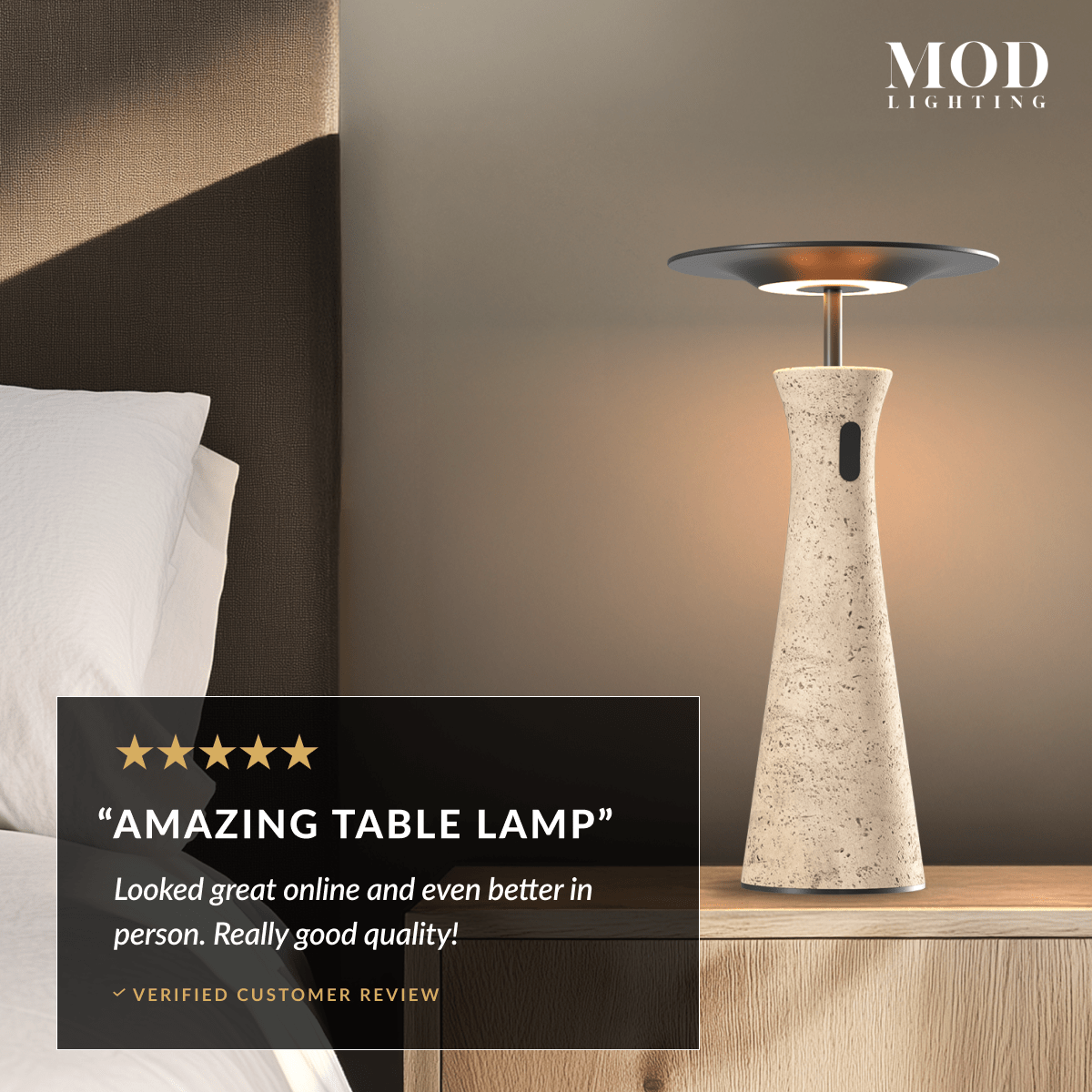 Dara tapered travertine table lamp with black disc shade beside a bed, glowing warm white