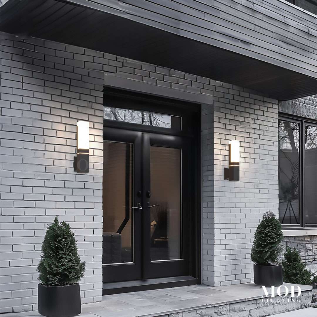 Falcon black outdoor sconces, slim dual-glow bars flanking a black glazed front door on grey brick at dusk