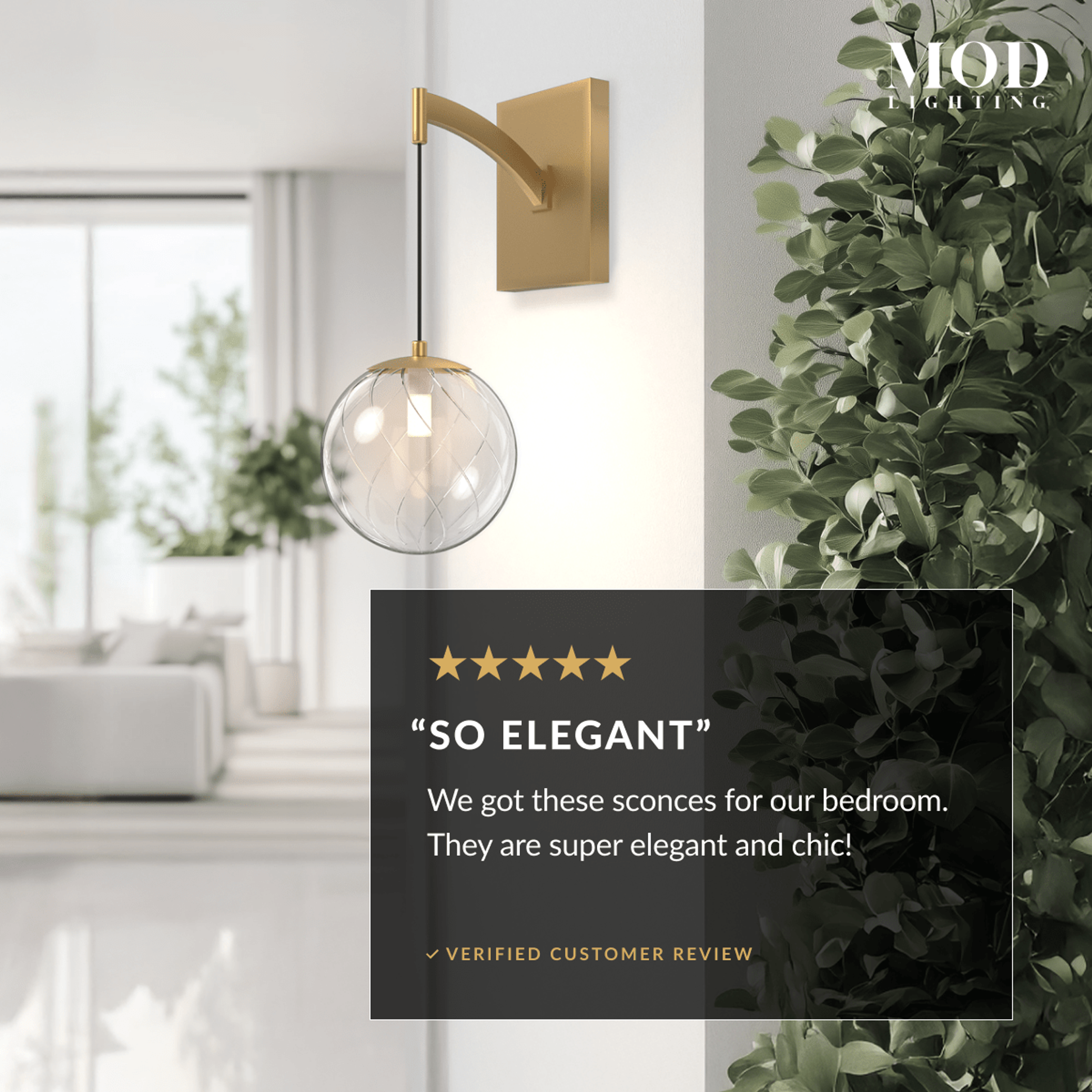 Mirella Sconce, brushed-gold arm and clear glass globe, mounted on a bright wall beside greenery