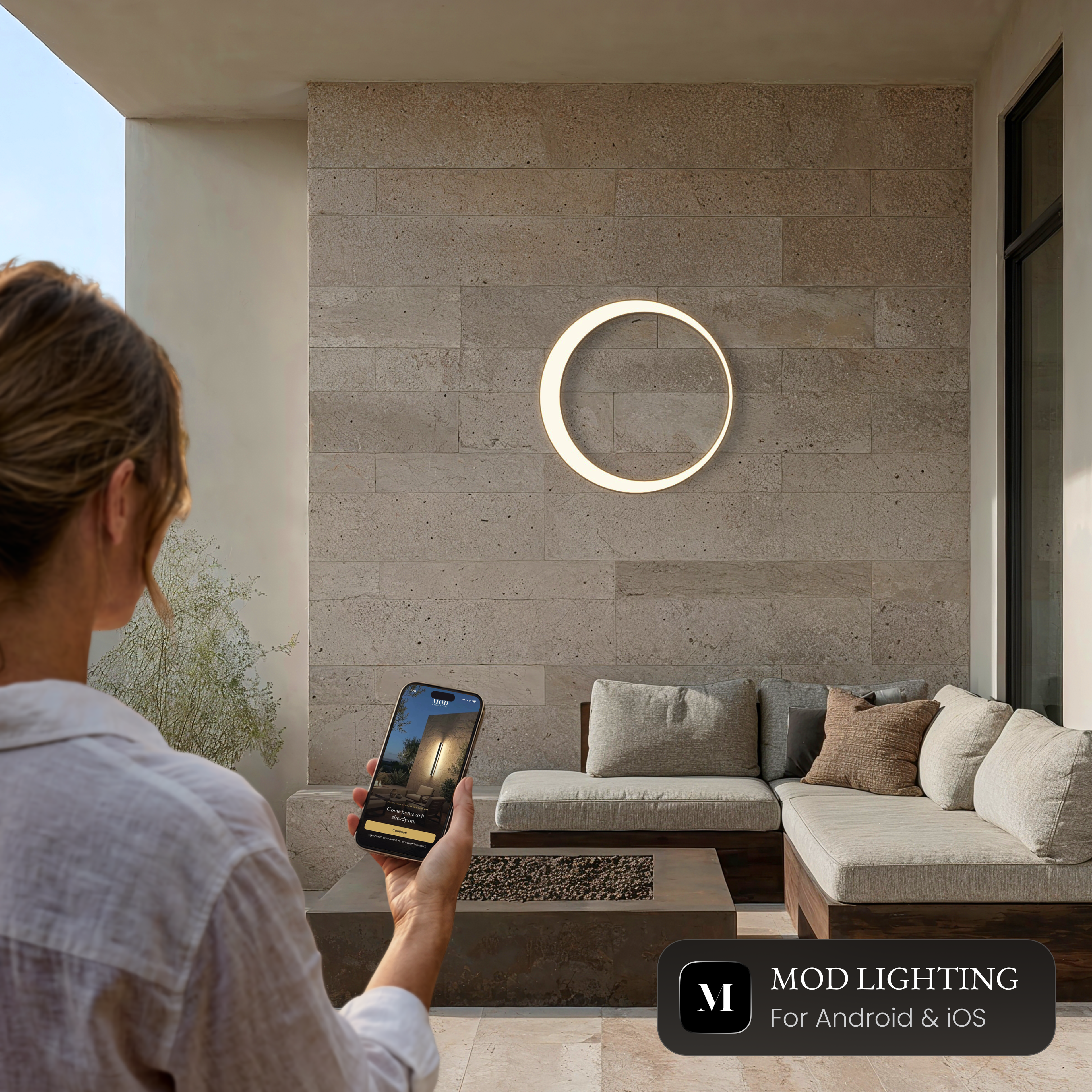 Gleam Smart round LED wall light on a stone wall, controlled from the MOD LIGHTING app on a phone