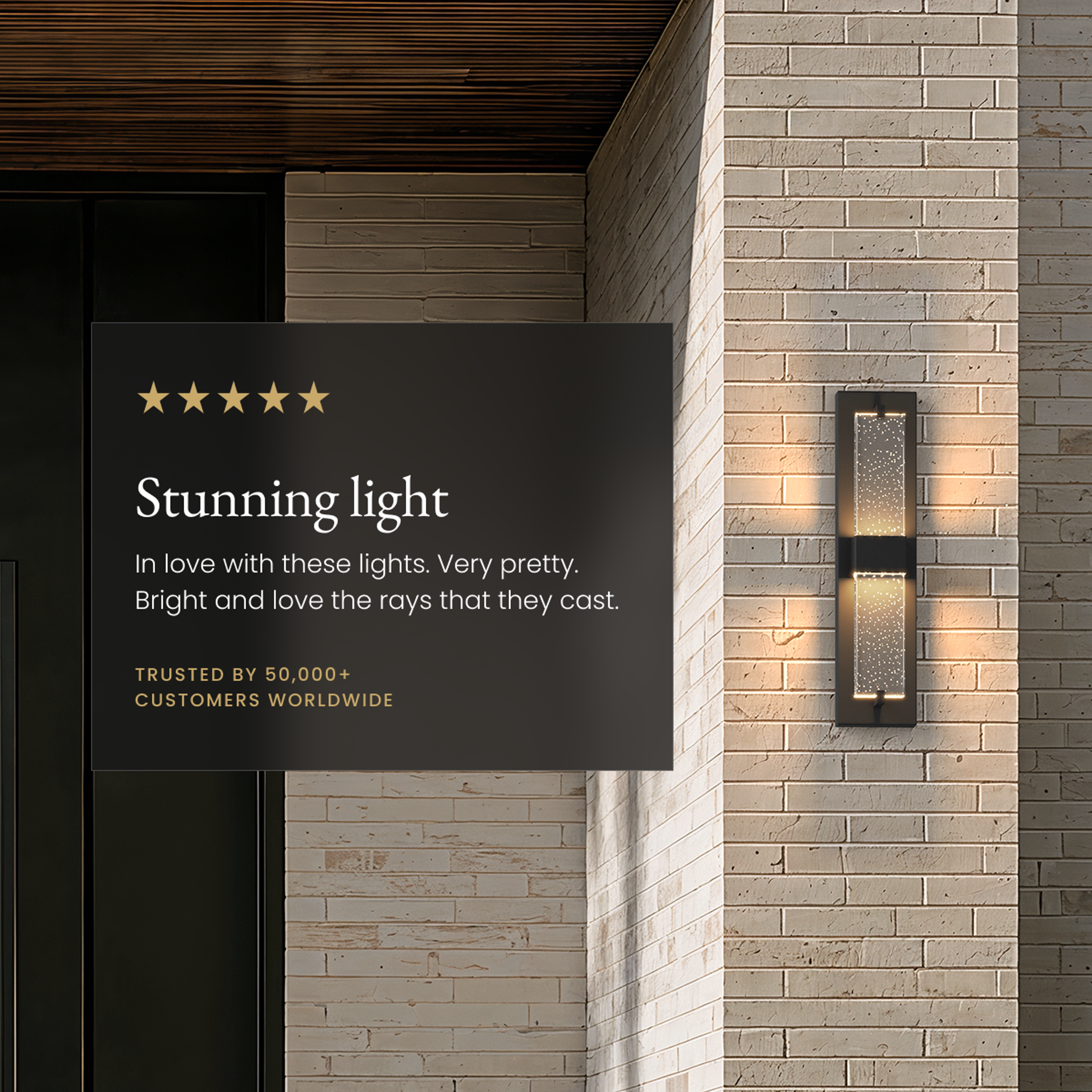 Noir black outdoor sconce, tall dual seeded-glass bars glowing on a stone-brick facade beside a five-star review card