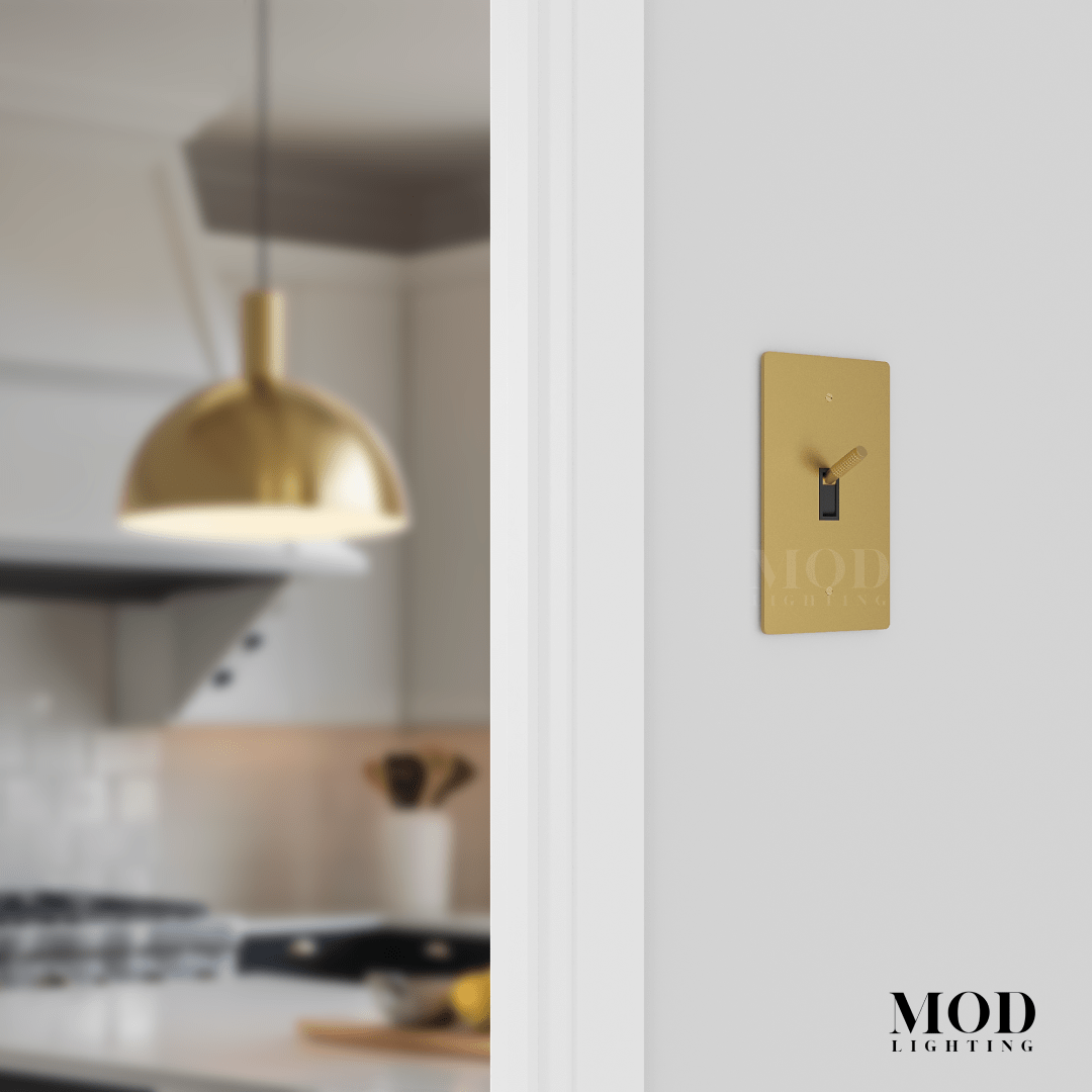 Modern Toggle Switch in brushed gold on a white wall beside a kitchen with a gold dome pendant