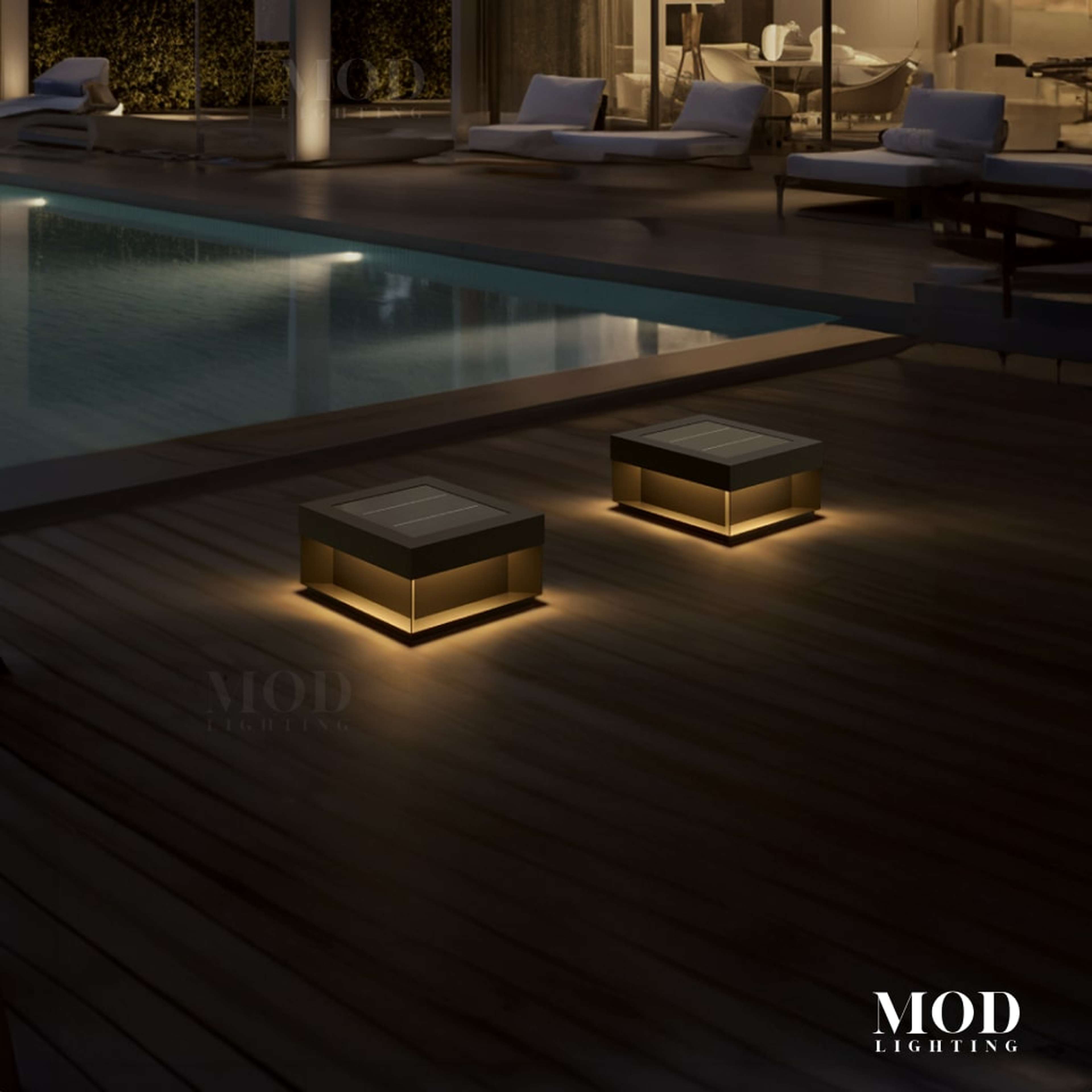 Chaz black square solar deck lights glowing warm on a wood pool deck beside a lit modern home at night