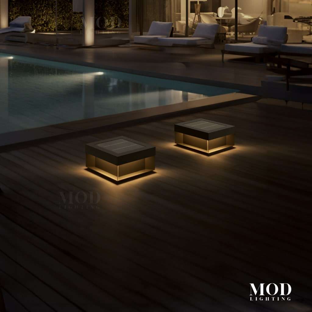 Chaz black square solar deck lights glowing warm on a wood pool deck beside a lit modern home at night
