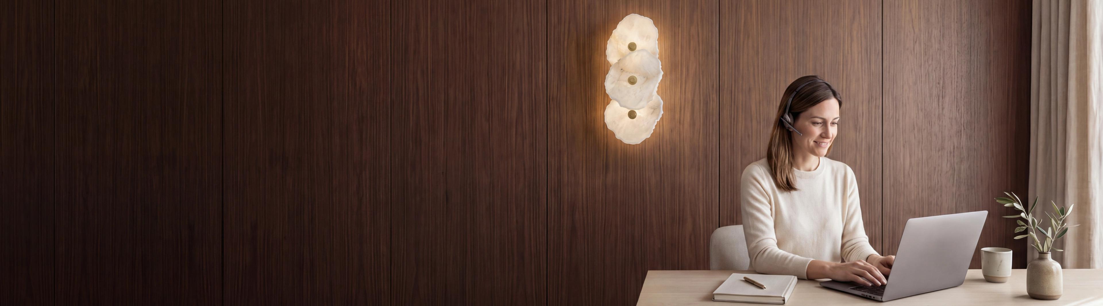 A MOD LIGHTING support specialist at a desk with a headset, beside an Alba alabaster sconce on a walnut wall
