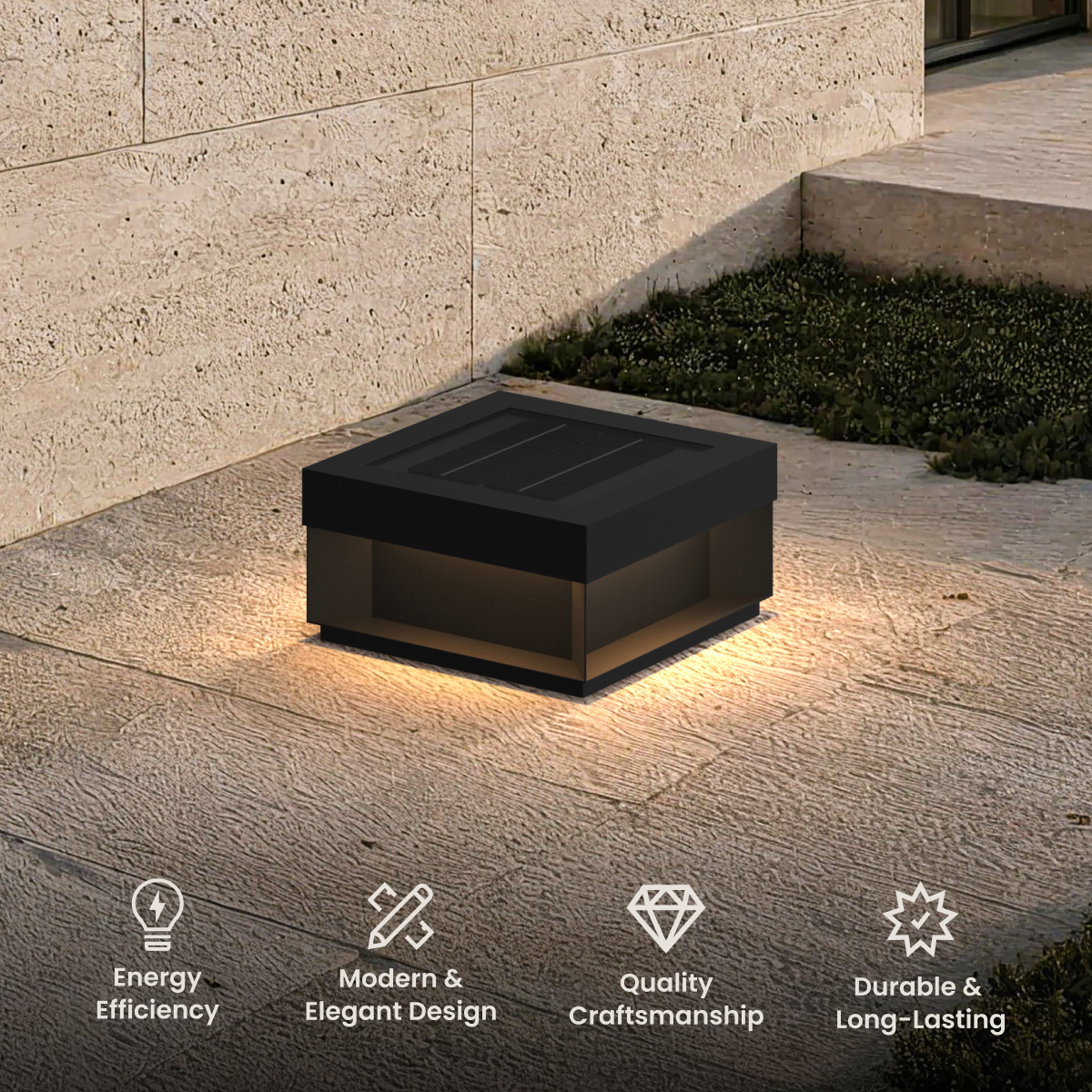 Chaz solar lamp, low square black bollard with warm glow, on a stone patio beside a wall