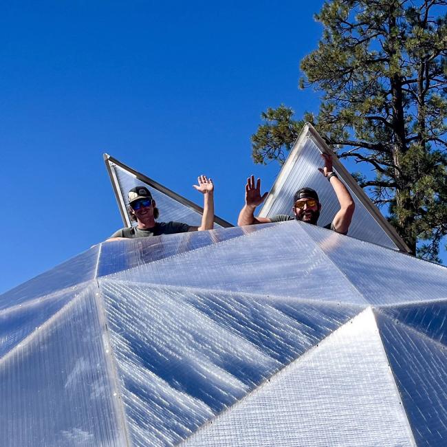 Two greenhouse builders standing in the top vents of the greenhouse waving and smiling
