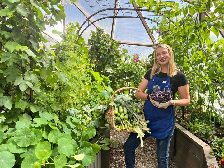 smiling woman holding a basket of produce in a lush greenhouse