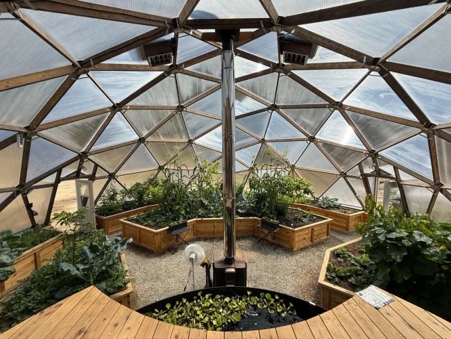 42′ Growing Dome greenhouse interior with pond deck, raised vegetable beds, wood-burning stove, and top-panel exhaust fans.