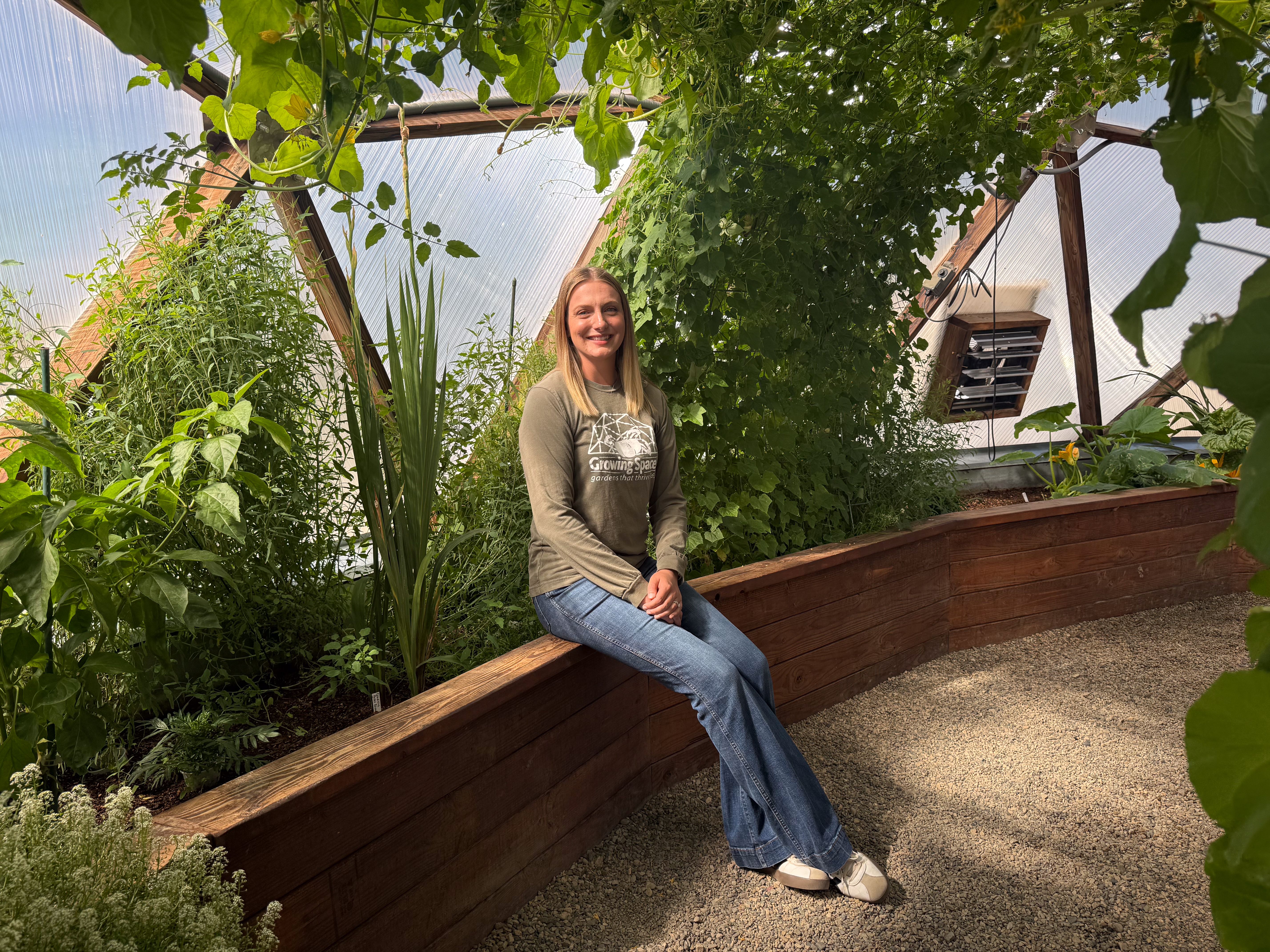 Person seated on the edge of a planted raised bed inside a greenhouse