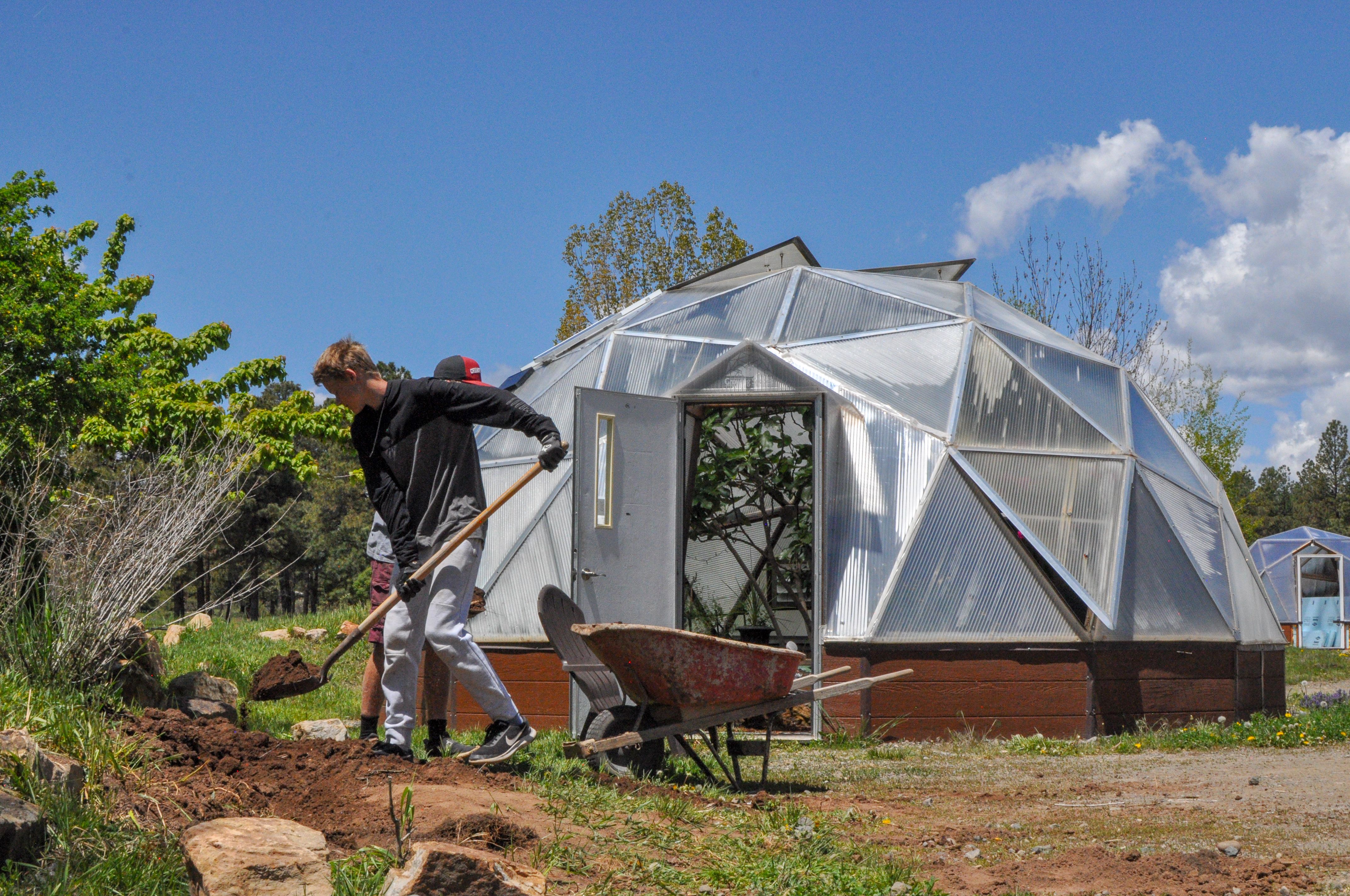 Two young men shoveling soil from a hillside into a wheelbarrow with a growing dome in background