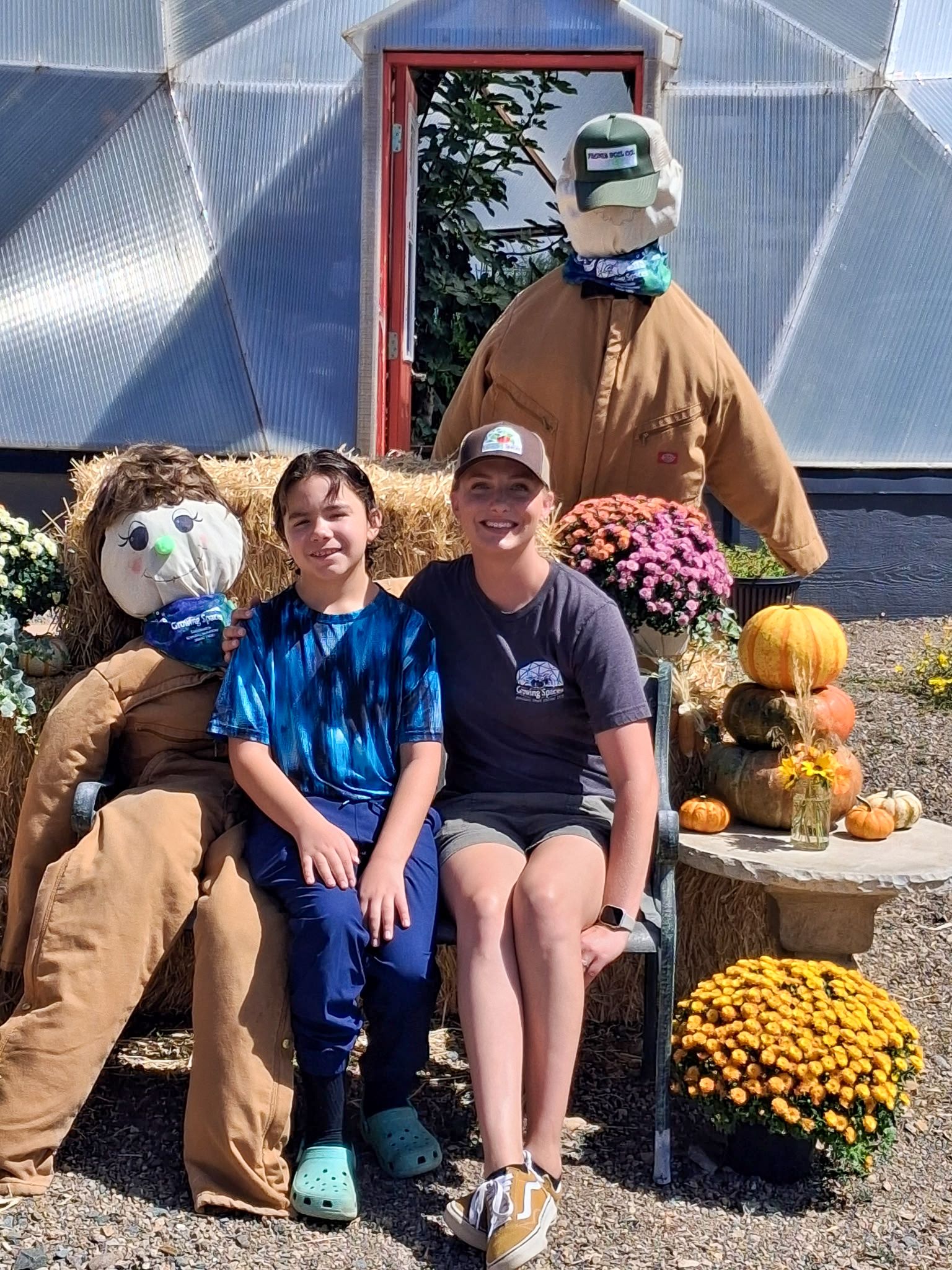 Two people posing for a photo on a bench with fall decor and scarecrows