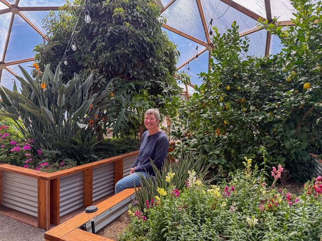 Woman seated on a garden bed in her Growing Dome greenhouse surrounded by large fruit trees and various flowers