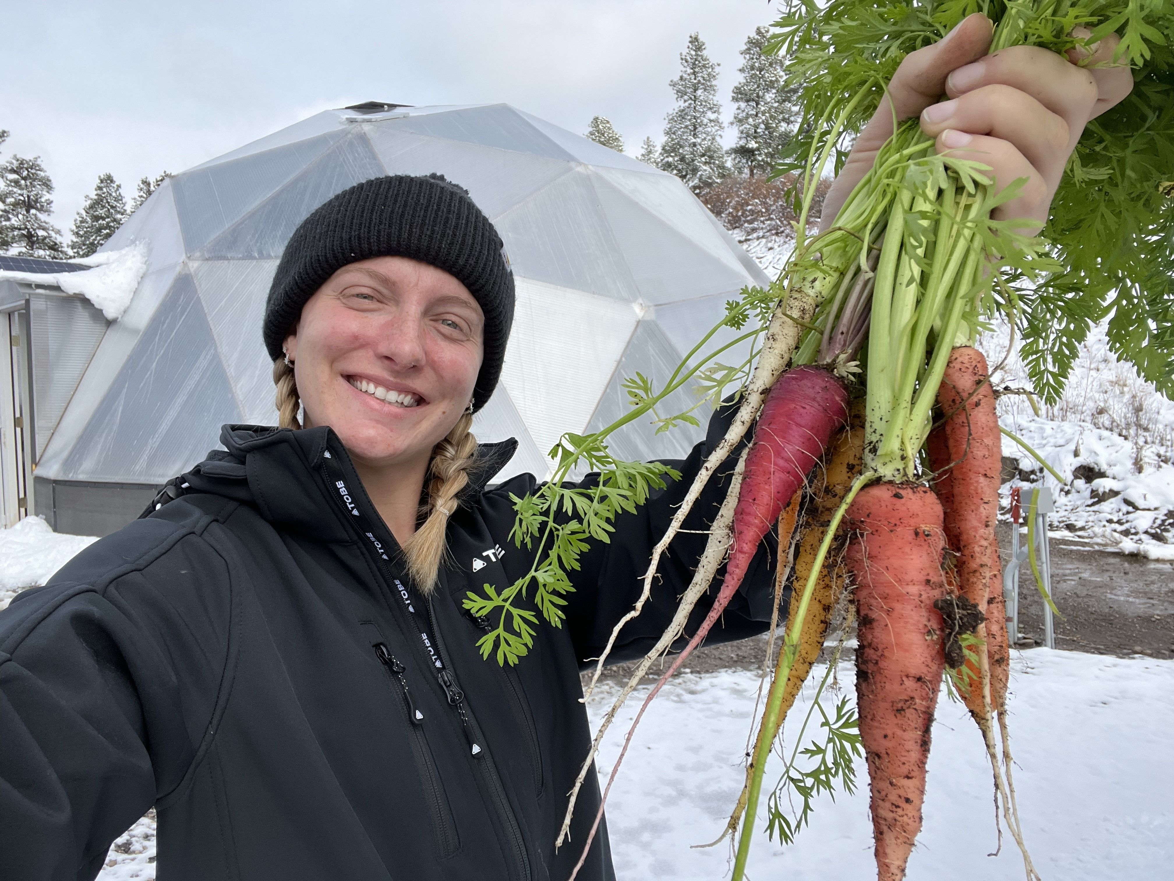 Woman in black jacket and beanie holding a handful of carrots with geodesic greenhouse and snow behind her