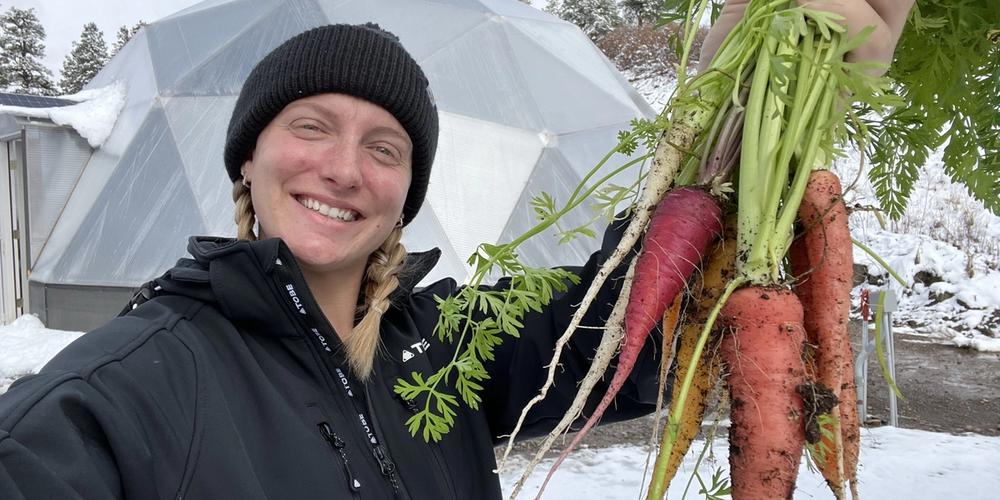Woman in black jacket and beanie holding a handful of carrots with geodesic greenhouse and snow behind her