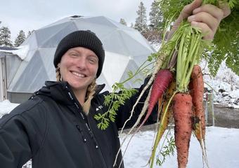 Woman in black jacket and beanie holding a handful of carrots with geodesic greenhouse and snow behind her