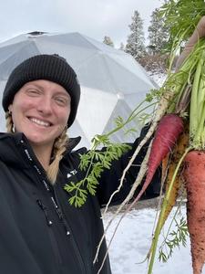 Woman in black jacket and beanie holding a handful of carrots with geodesic greenhouse and snow behind her