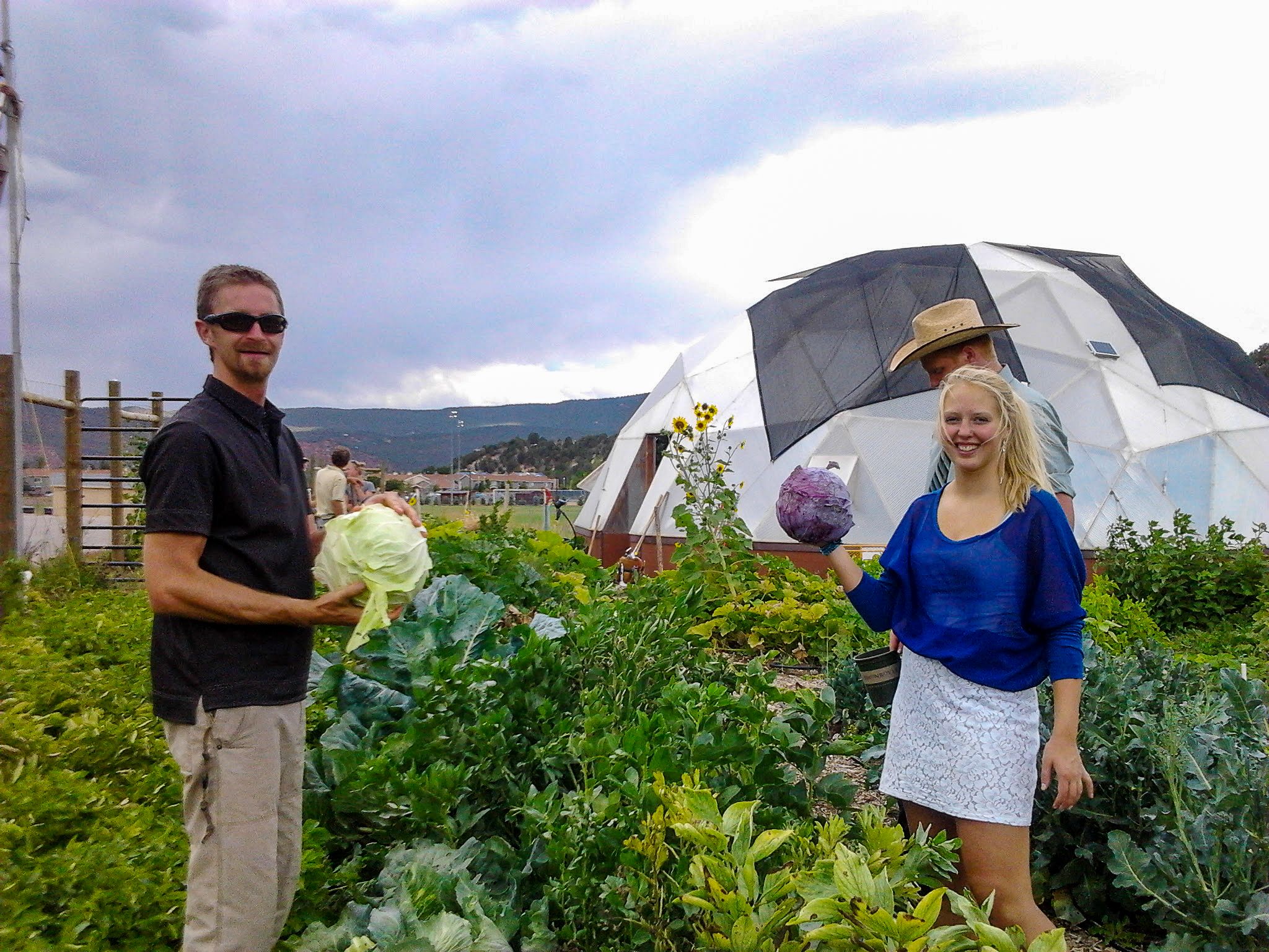 people showing off cabbages they harvested outside with a geodesic growing dome greenhouse in the background
