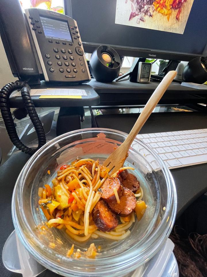 a bowl of spaghetti, sausage, and leftover ratatouille in a clear bowl with a bamboo fork sticking out on an office desk