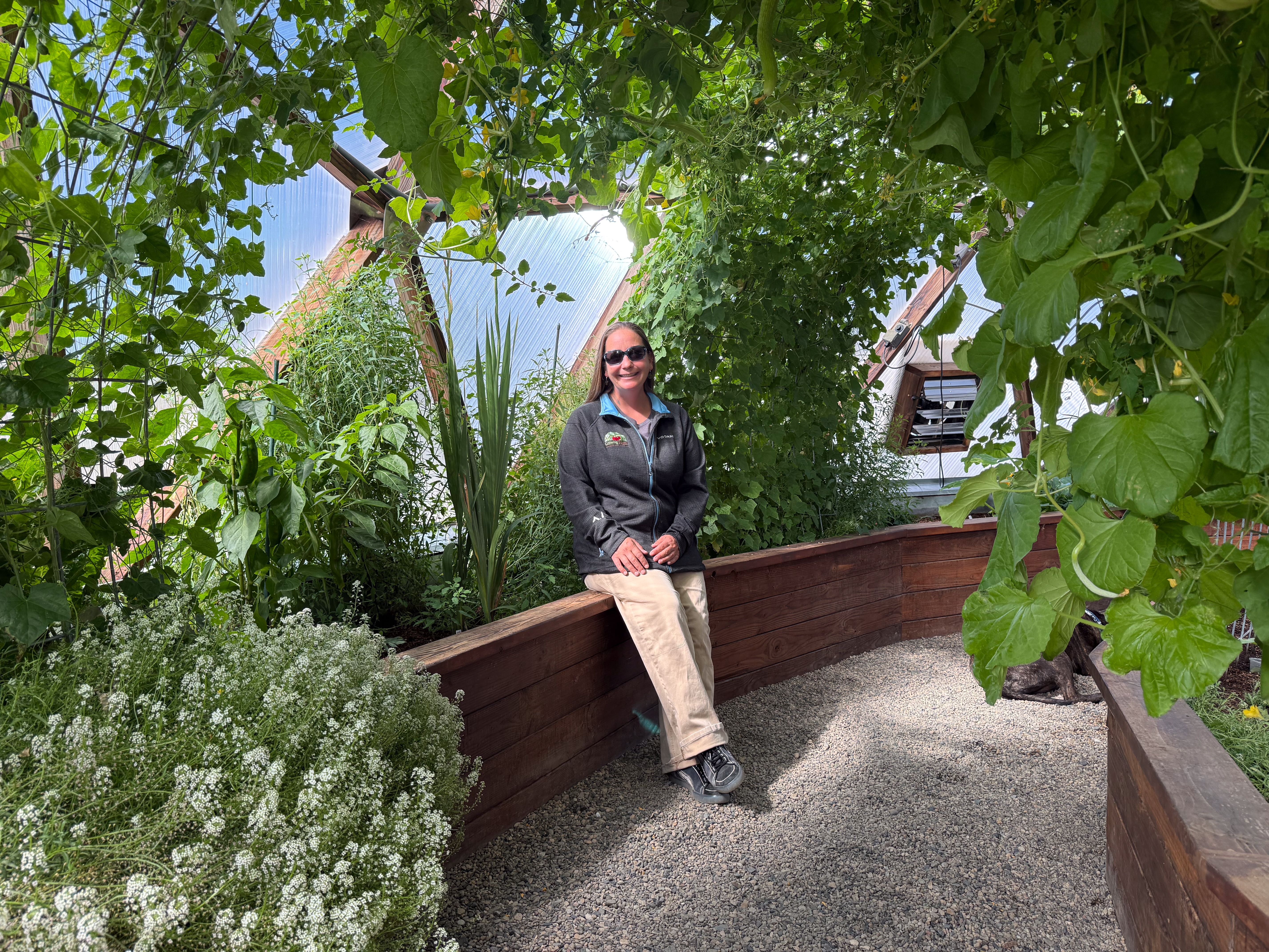 Person in a gray jacket seated beside plants inside a greenhouse