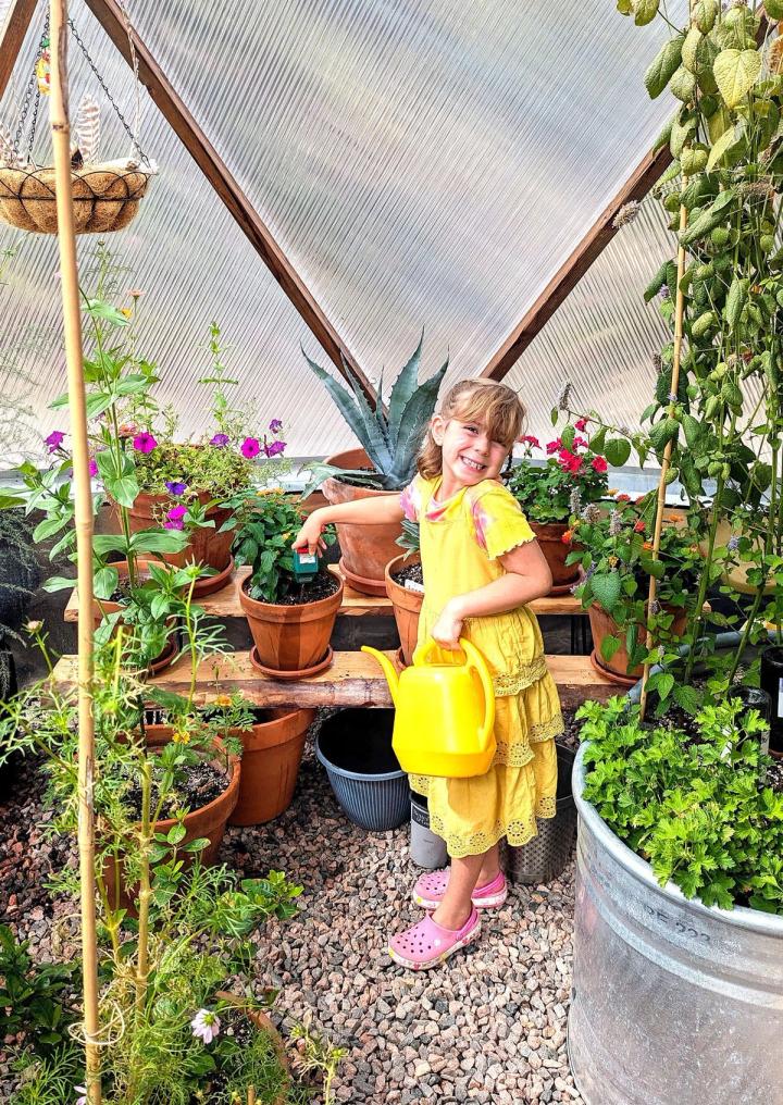 A young lady in a yellow dress is watering plants inside a Growing Dome. In one hand, she holds a yellow watering can. With the other, she places a moisture meter into the soil of a clay pot.