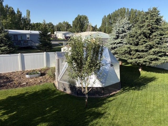 Backyard greenhouse surrounded by manicured lawn, small trees, and a white fence