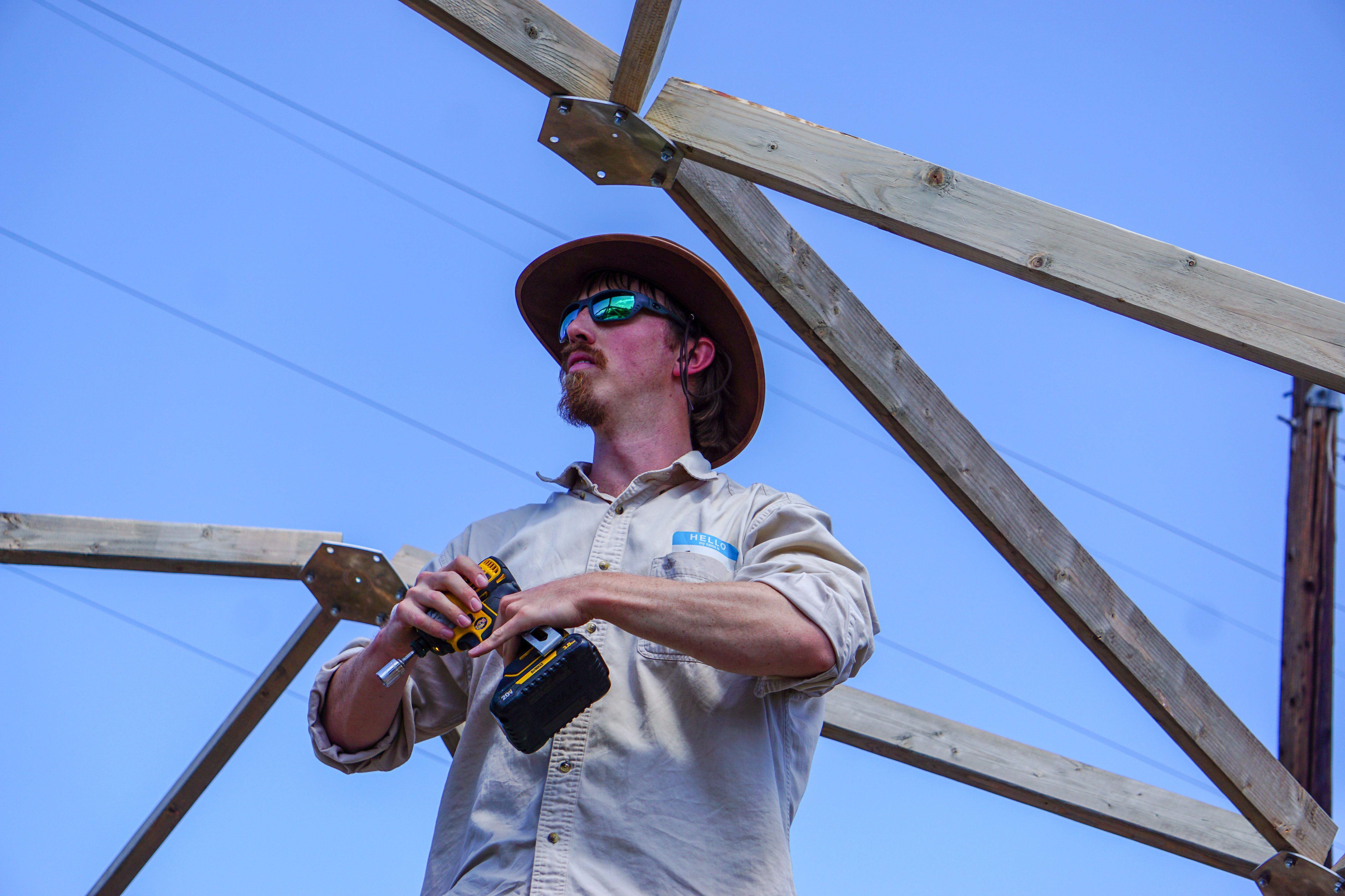 Man in a hat and sunglasses holding a drill with a geodesic frame behind him