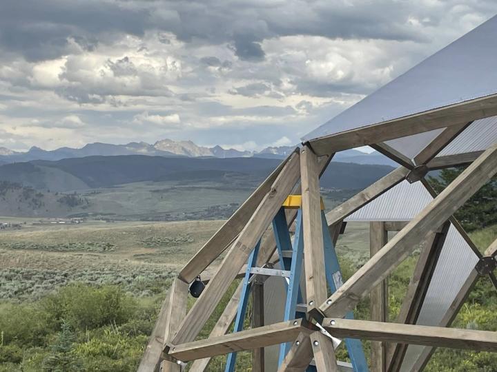 Growing dome under construction with a ladder inside, exposed struts, and scenic mountain views behind