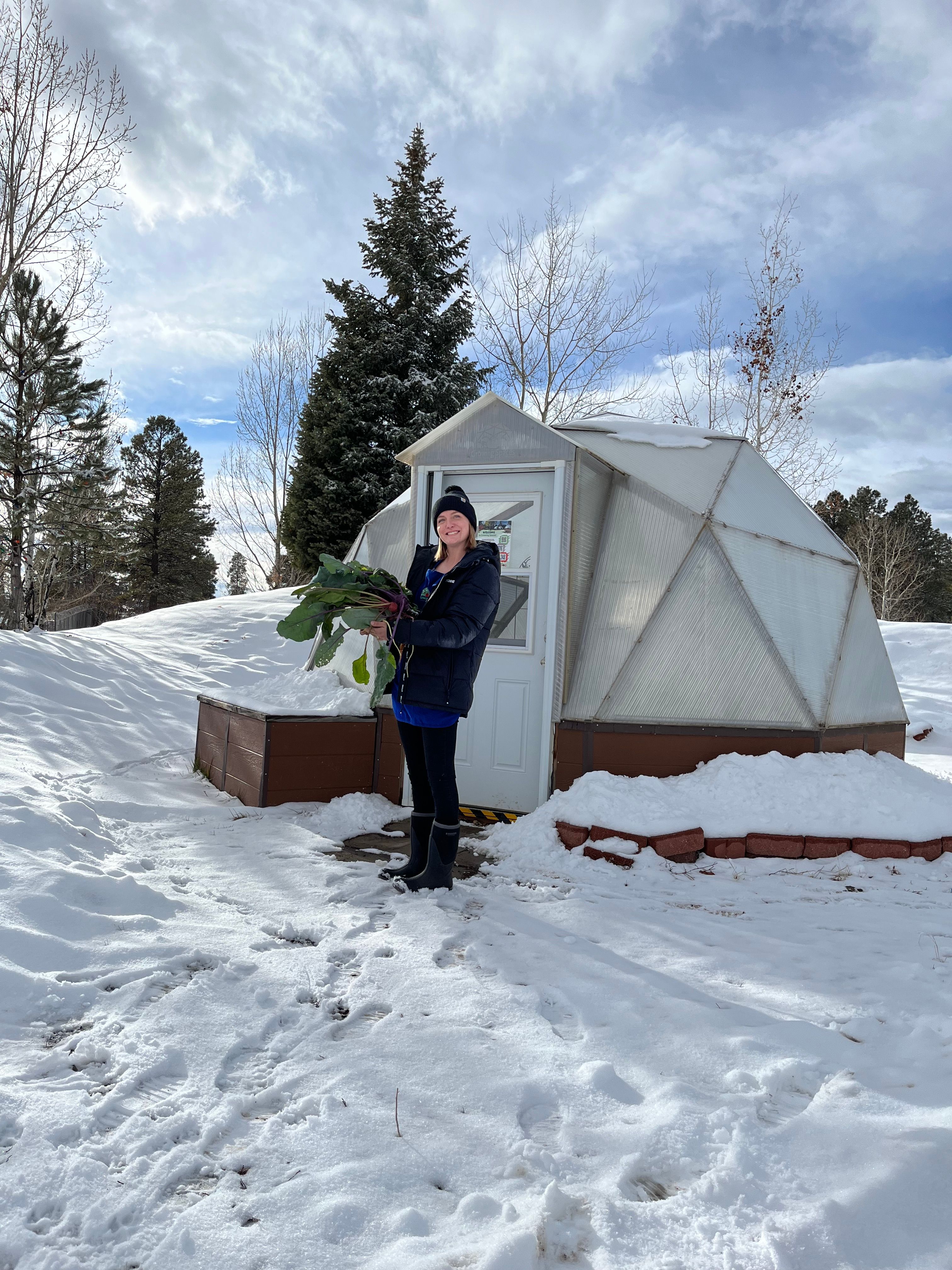 Woman holding a large basket of produce standing outside a greenhouse with snow on the ground