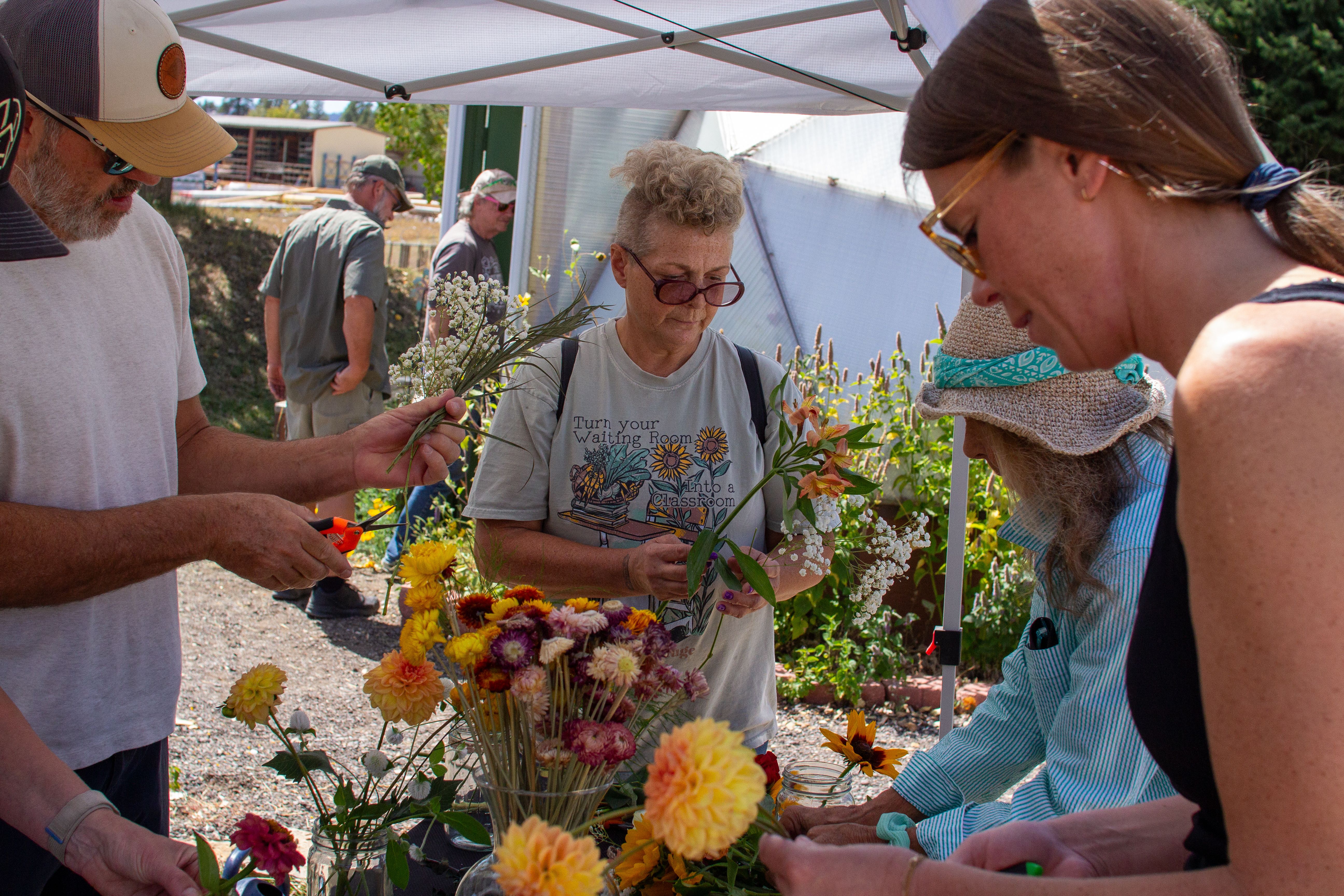 Group of people gathered around a table making flower arrangements with two others entering a greenhouse in the background