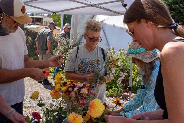 Group of people gathered around a table making flower arrangements with two others entering a greenhouse in the background