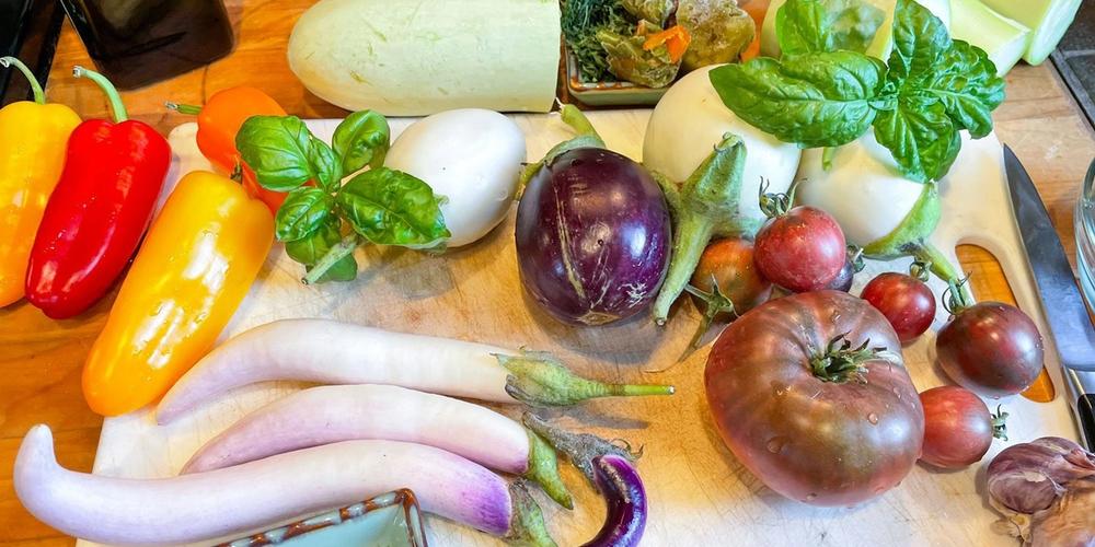 various raw ingredients on a cutting board ready to make ratatouille: fresh tomatoes, eggplant, herbs, peppers, and zucchini
