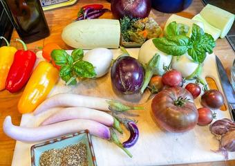 various raw ingredients on a cutting board ready to make ratatouille: fresh tomatoes, eggplant, herbs, peppers, and zucchini