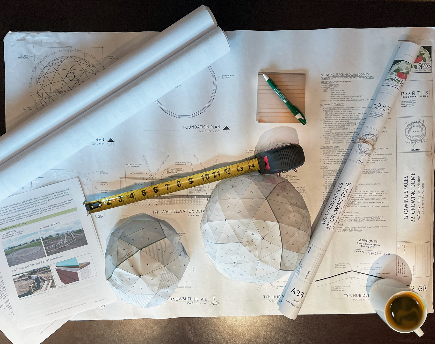 Greenhouse plans on a table with a tape measure, coffee cup, and notepad
