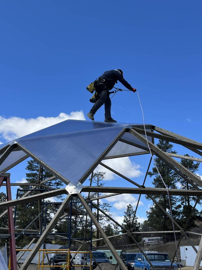 Man on top of a geodesic greenhouse during construction tied off with a rope and harness