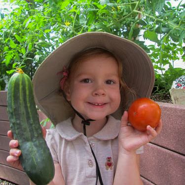 Child in a sun hat holding up a cucumber and a tomato