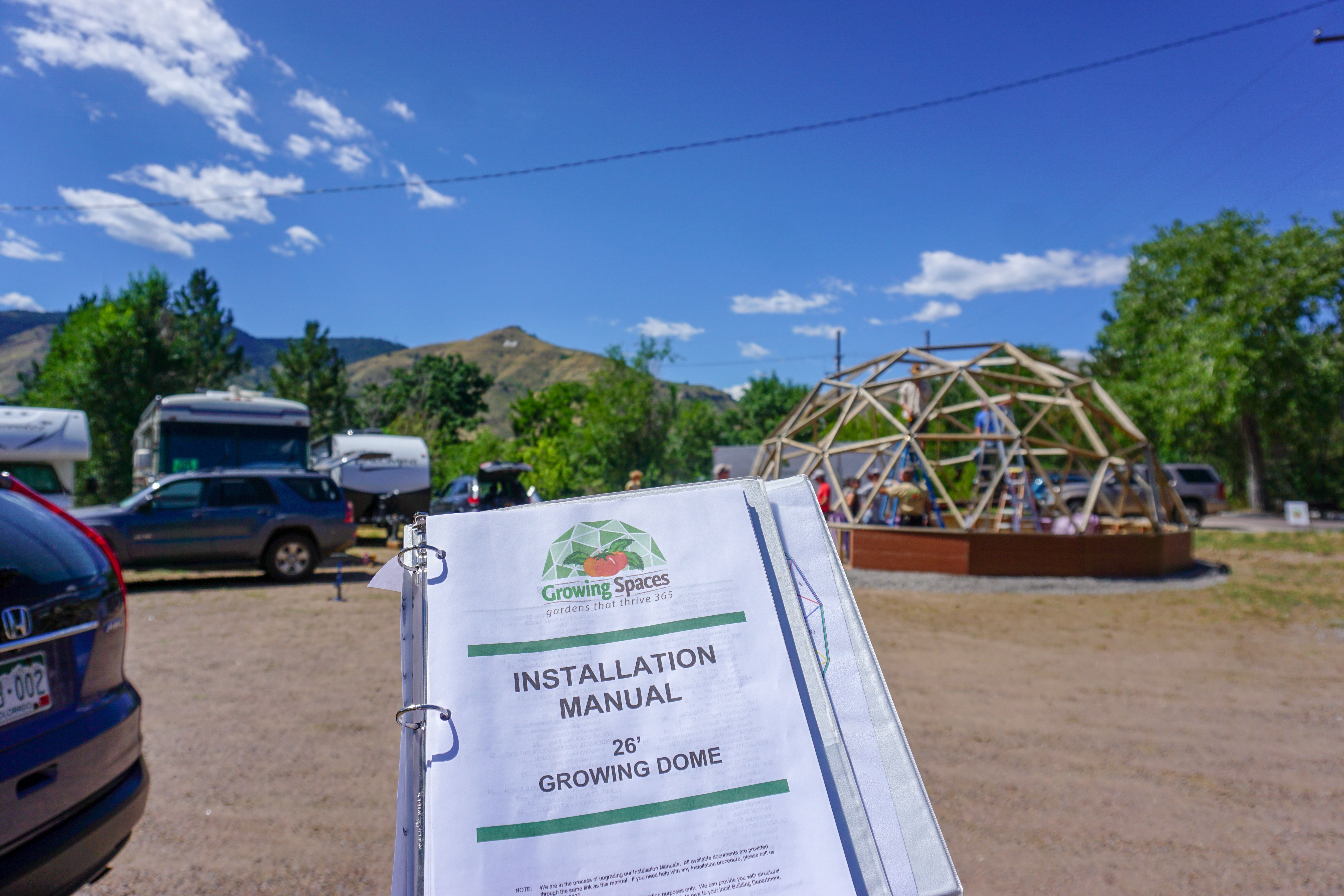 in the foreground an installation manual for a 26' Growing Dome greenhouse in the background the framework of the greenhouse has been installed