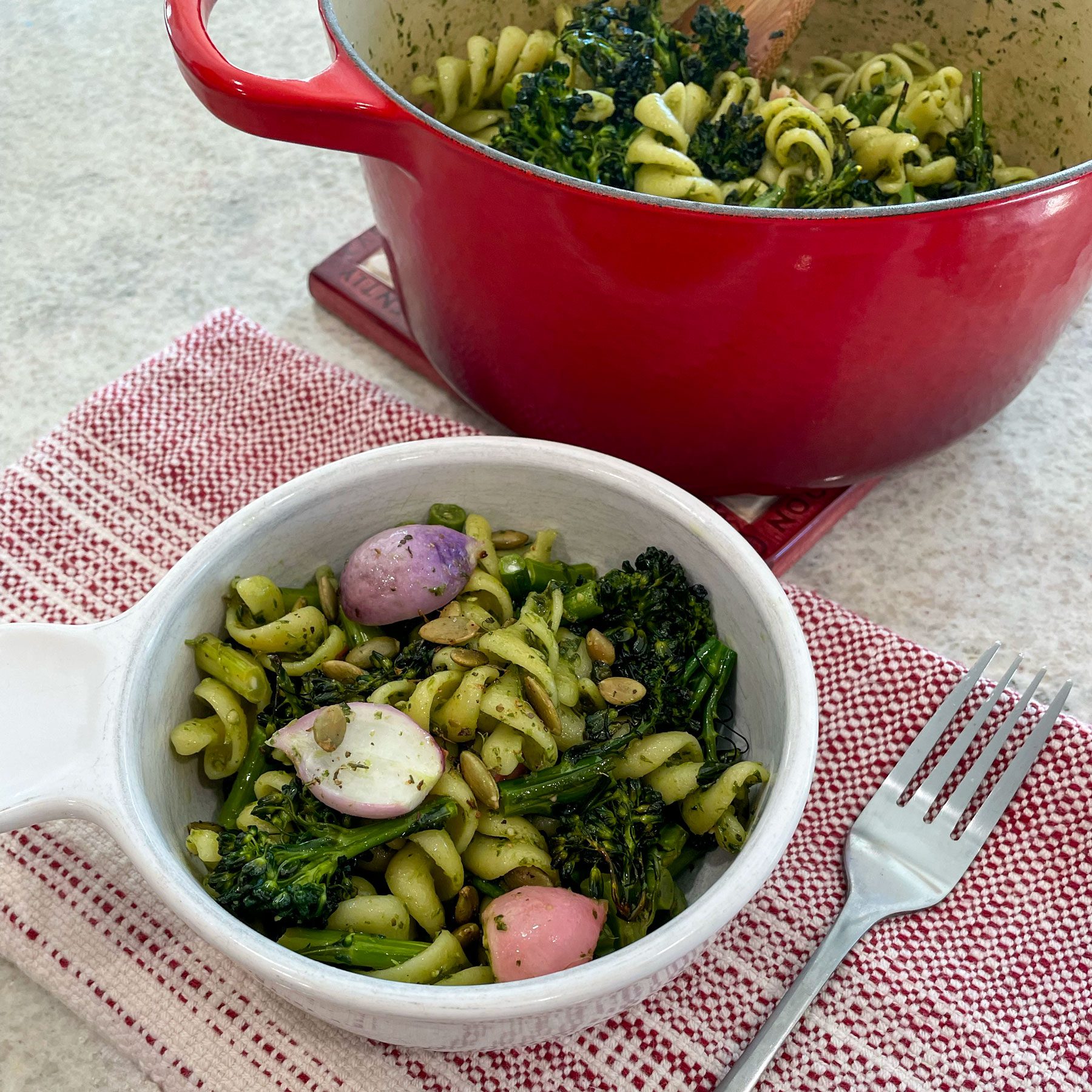 bowl of winter pesto pasta salad served up in front of the pot filled with pasta broccoli and radishes