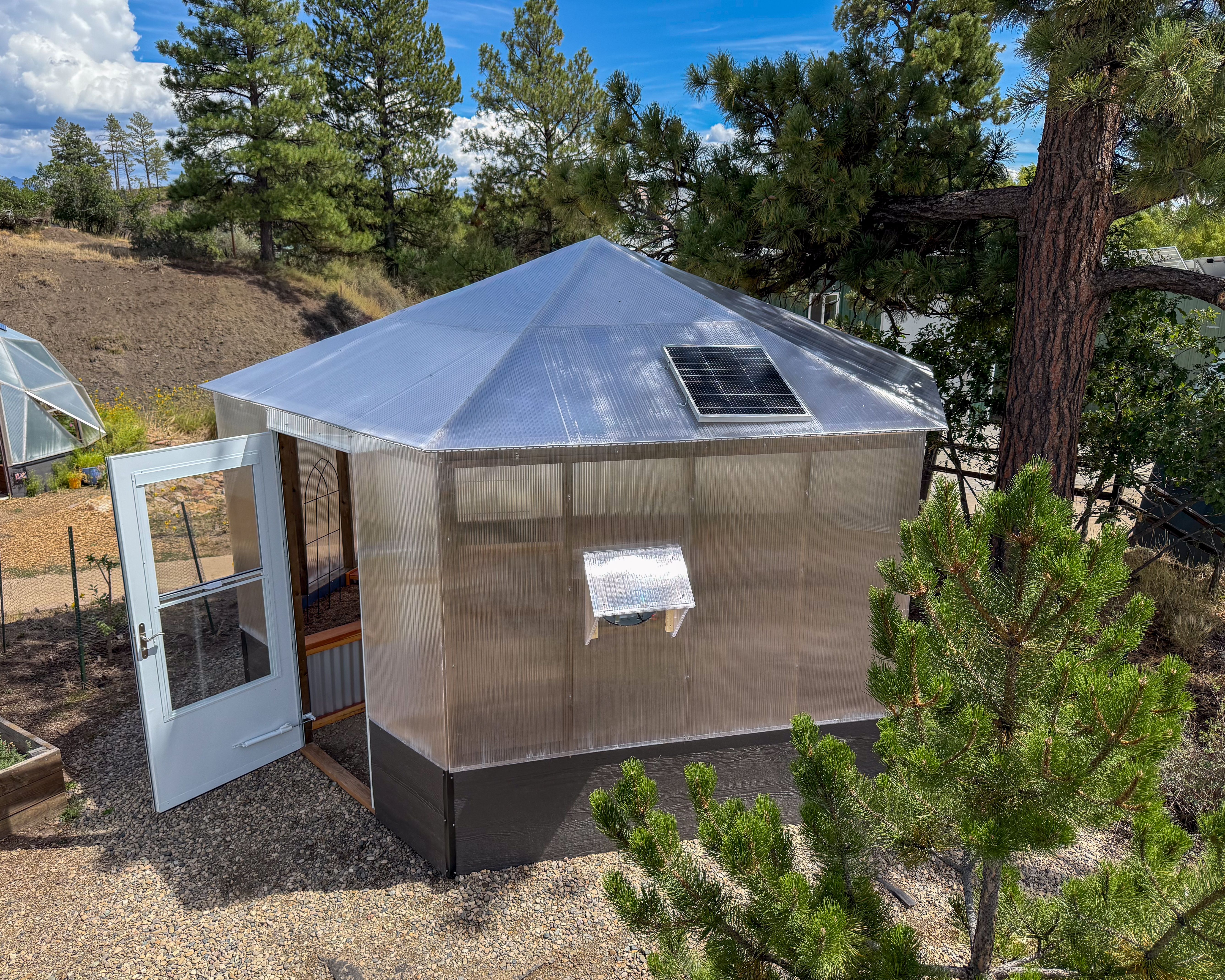 Exterior of a Growzebo backyard greenhouse with an open white front door and solar powered fan