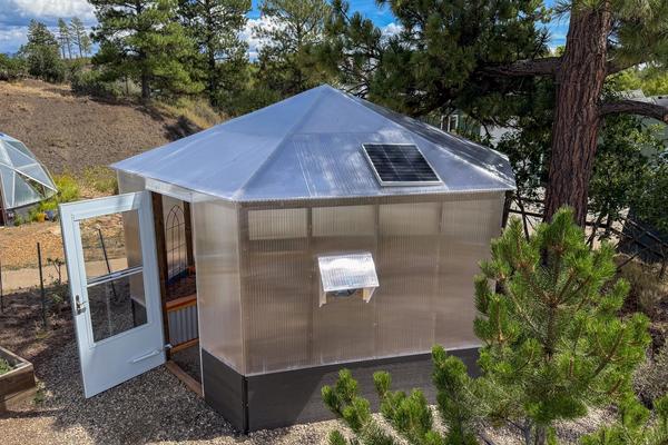 Exterior of a Growzebo backyard greenhouse with an open white front door and solar powered fan