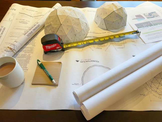 Greenhouse plans and installation instructions laid out on a desk with a tape measure, notepad, pen, and cup of coffee.