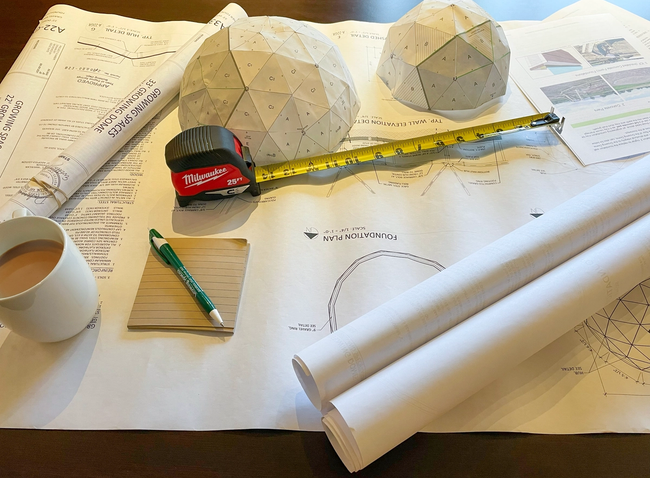 building plans paper dome models notes tape measure and coffee on a desk