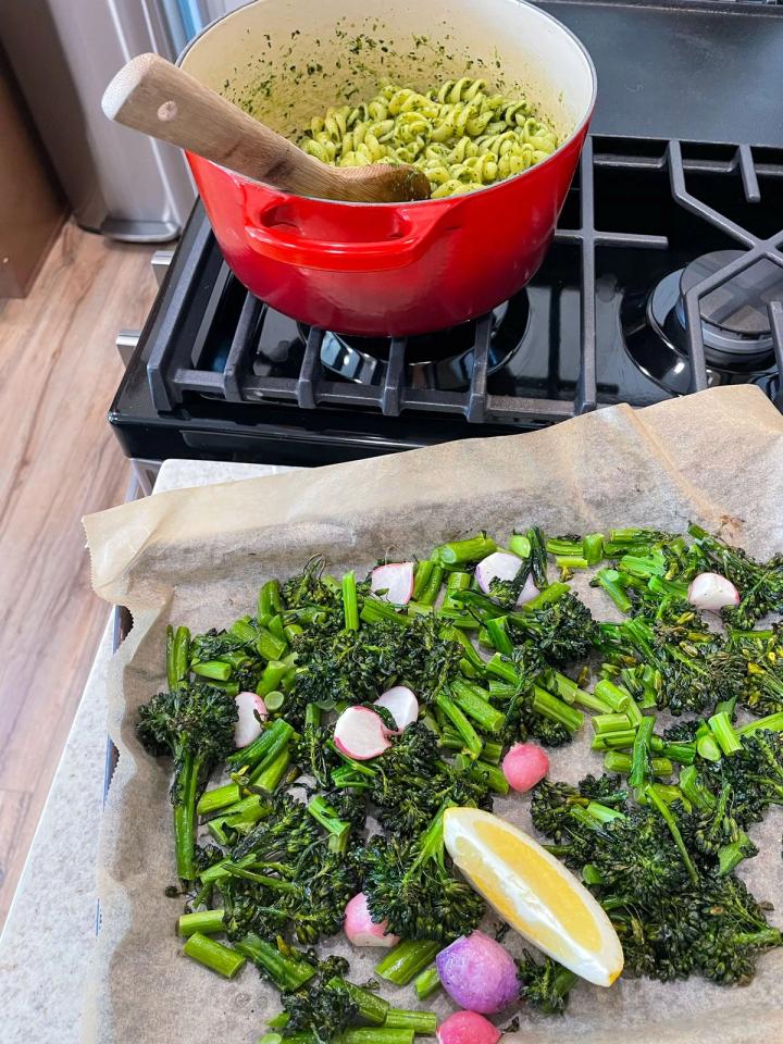 pot of pasta mixed with pesto on the stove with a tray of roasted radishes and broccolini in the foreground with a wedge of lemon