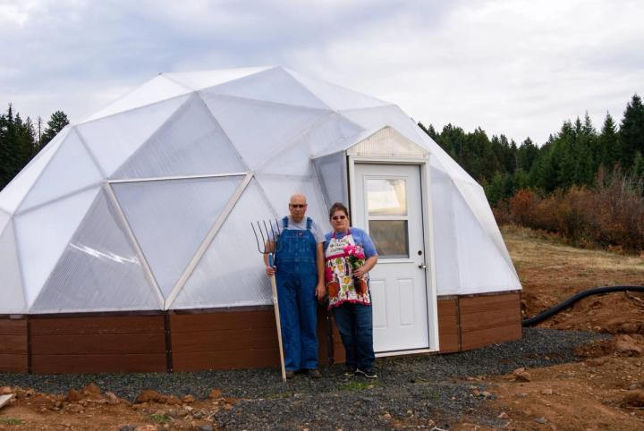 Paula and Keith Rigby's doing an American Gothic pose in front of their Backyard Greenhouse in Idaho