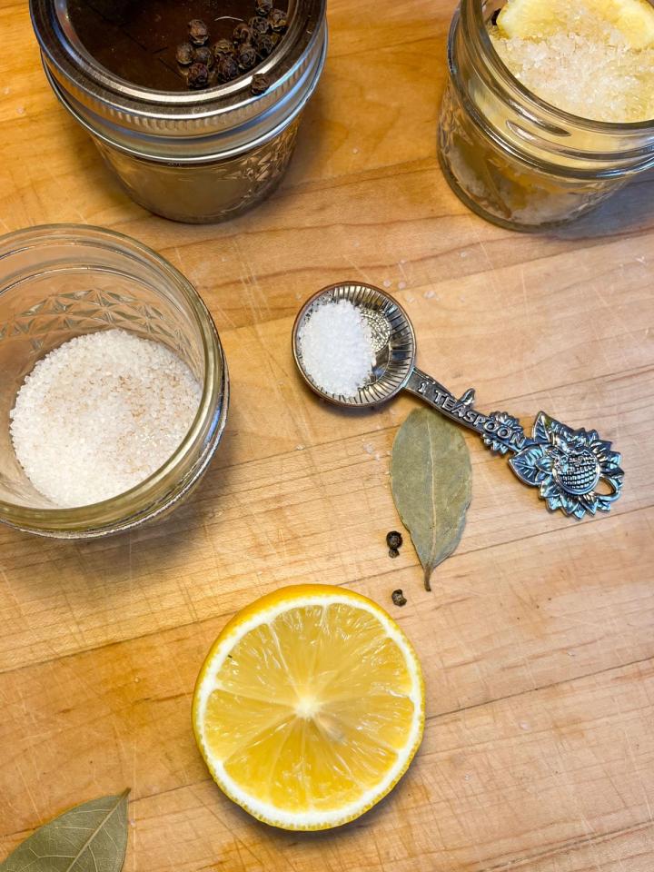 the ingredients for preserved lemons on a wooden butcher block: lemons, bay leaves, peppercorns, a teaspoon filled with salt and glass jars