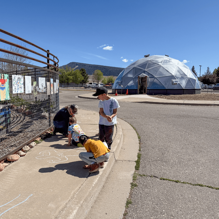 Students making shadows on the sidewalk and tracing them with chalk with a growing dome in the background