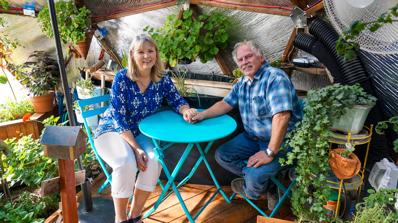 two people sitting in their greenhouse holding hands and smiling at the camera