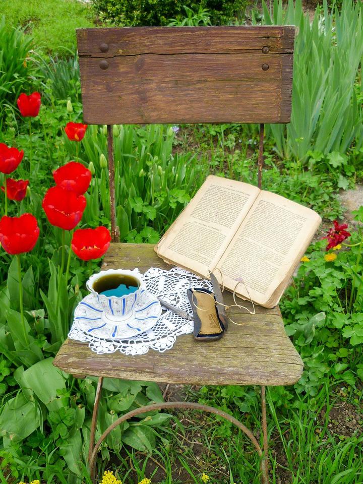 Weathered wooden chair sitting outside in a garden with red tulips blooming beside it. There is a book, tea cup, doily, and a pair of glasses sitting on the chair.